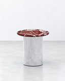 Orbitae side table featuring a circular burgundy veined stone top supported by a cylindrical silver-toned pedestal. The contrasting stone pattern and reflective base create a refined contemporary accent.