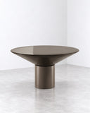 Silo dining table featuring a broad, softly contoured dark tabletop and a substantial cylindrical pedestal base. Its monolithic silhouette and smooth finish create a bold contemporary centerpiece.