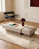 THERRA coffee table featuring a textured light travertine top and block-like stone supports arranged in an asymmetric composition. Its tactile natural surface and monolithic form bring warmth to contemporary interiors.