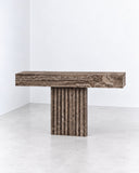 Therra console table featuring a long rectangular travertine top and a substantial vertically fluted stone pedestal. The natural porous texture and architectural proportions emphasize the sculptural character of the stone.