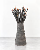 Sandrine Herlin Baobab Noir Small Lighting features a compact, tapered charcoal body with layered surface textures, perforated detailing, and branching stems accented with orange tips. Its sculptural silhouette evokes a stylised tree, creating a distinctive decorative lighting piece.