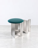 Sketch side table featuring a rounded teal tabletop supported by a sculptural polished metal base with curved vertical elements. The contrast between the saturated surface and reflective supports creates a distinctive contemporary accent.