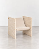 Adri lounge chair with a pale travertine frame, angular integrated armrests, and a neutral upholstered seat and backrest. Its geometric form highlights the tactile contrast between natural stone and fabric.