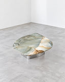 Round Oni coffee table featuring an organically shaped, richly veined stone top with green and amber tones and polished cylindrical metal supports. Its sculptural silhouette showcases the natural variation of stone in contemporary furniture design.