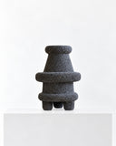 LS-02 volcanic rock sculpture featuring a stacked composition of rounded and flared forms above a short sculptural base. The coarse dark stone surface emphasizes the raw texture and distinctive geometry of the object.
