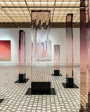 Group of vertical gradient sculptures displayed in a minimalist gallery with garden view
